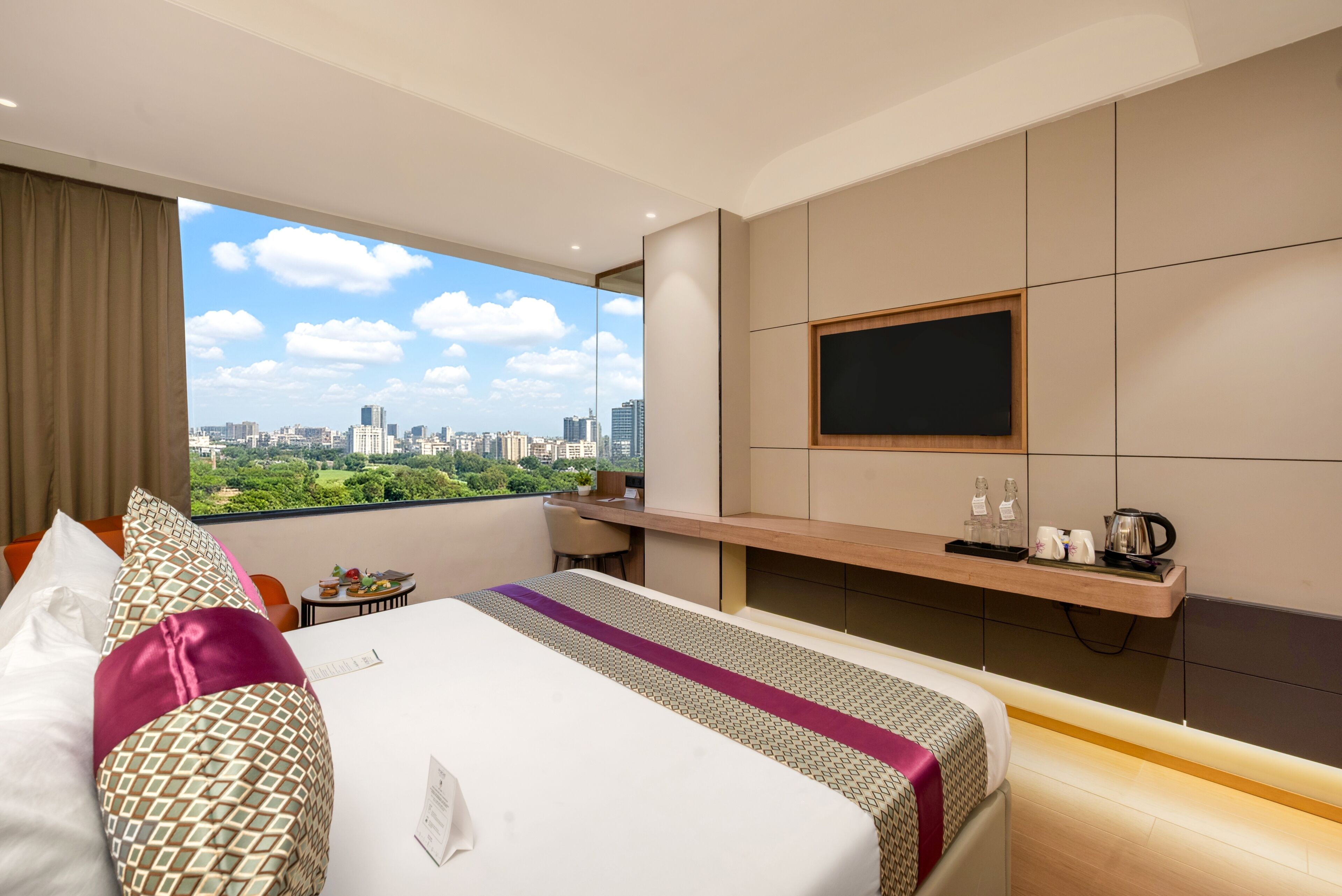A hotel room with a large bed, a TV mounted on the wall, a desk with a chair, and a view of the cityscape.