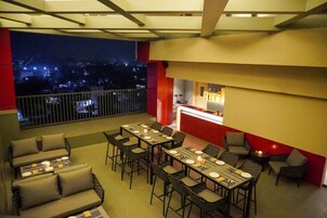 Un comedor con mesas dispuestas para cuatro personas, una zona de bar y un balcón con vista a la ciudad de noche.