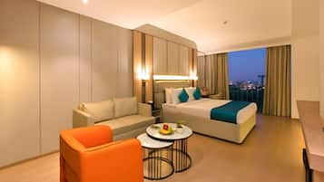 Una habitación de hotel moderna con una cama, un sofá, una mesa de centro y vistas a la ciudad al anochecer.