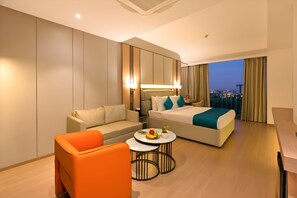 Una habitación de hotel moderna con una cama, un sofá, una mesa de centro y vistas a la ciudad al anochecer.