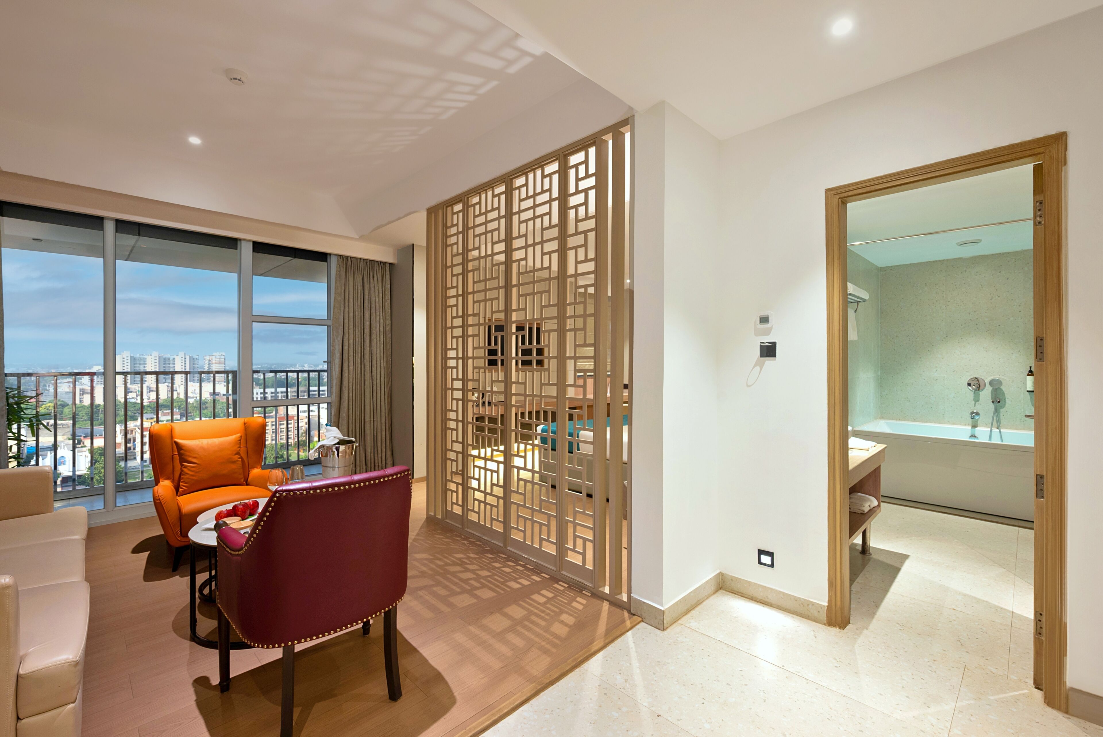 A modern hotel room with a city view, a seating area with a red chair, and a separate bathroom.