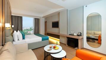 A hotel room with a bed, a sofa, a dining table with fruit, a coffee station, and a TV.