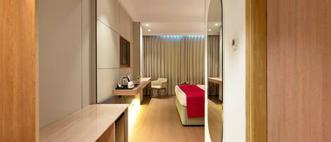 A hotel room with a wooden desk, a bed with a red blanket, a mirror, and a view of the room through a door.