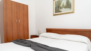 A hotel room with a wooden wardrobe, a bed with a white comforter and a gray blanket, a nightstand with a lamp, and a framed painting of a forest scene on the wall.