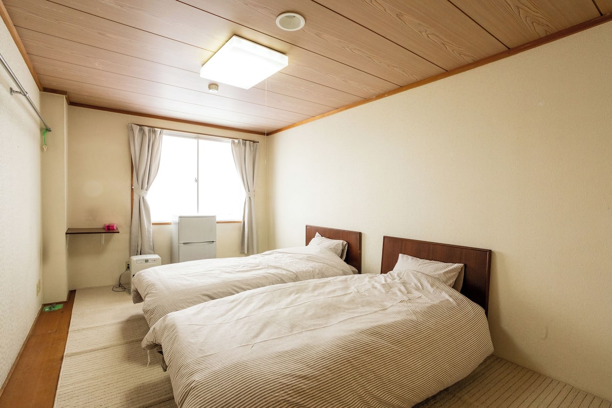 A room with two beds, a window with curtains, a desk, and a ceiling light.