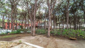 A forested area with a gravel path, a small garden, and a building with a red facade in the background.