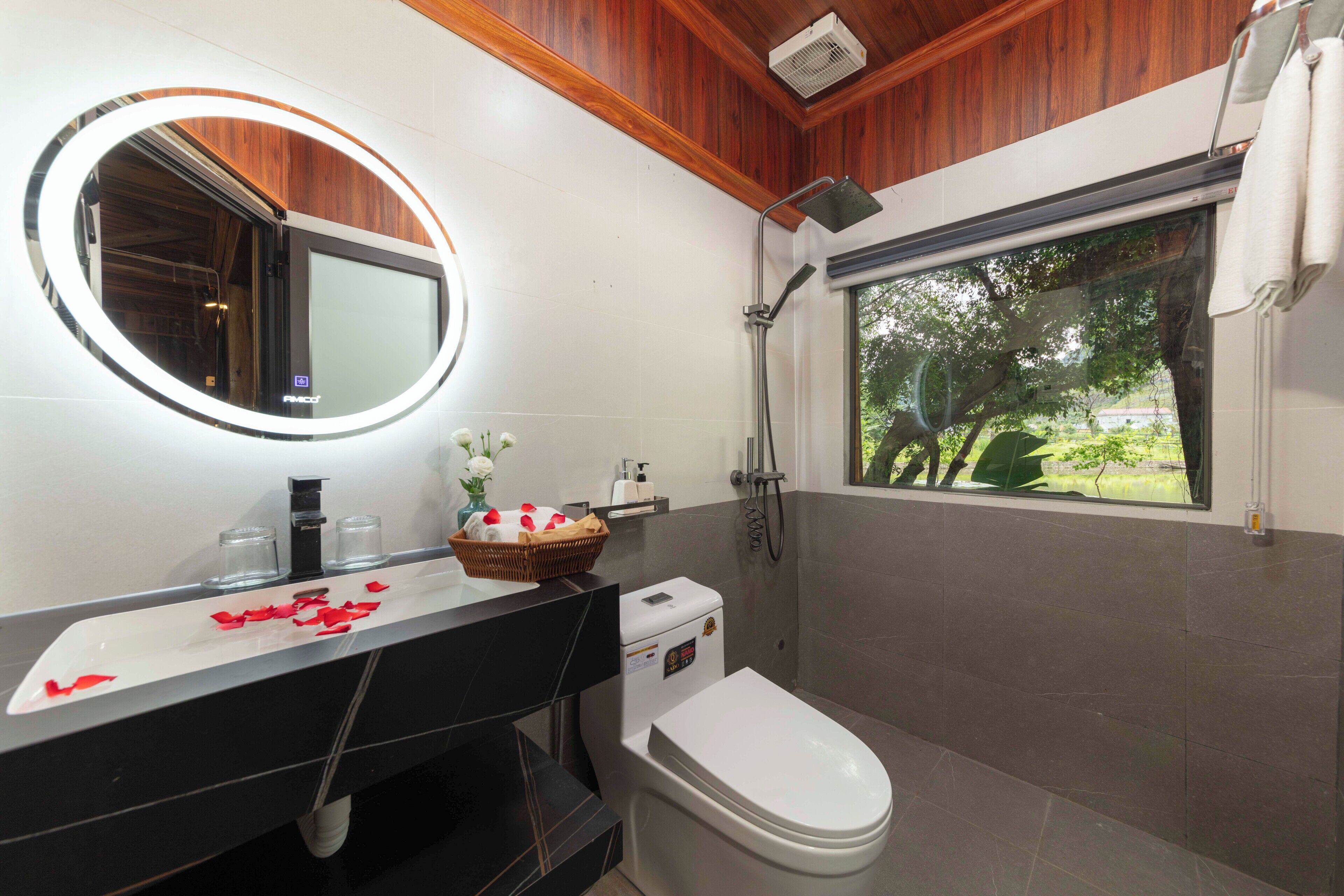 A bathroom with a wooden ceiling, a white sink with a mirror, a white toilet, and a shower with a view of trees.