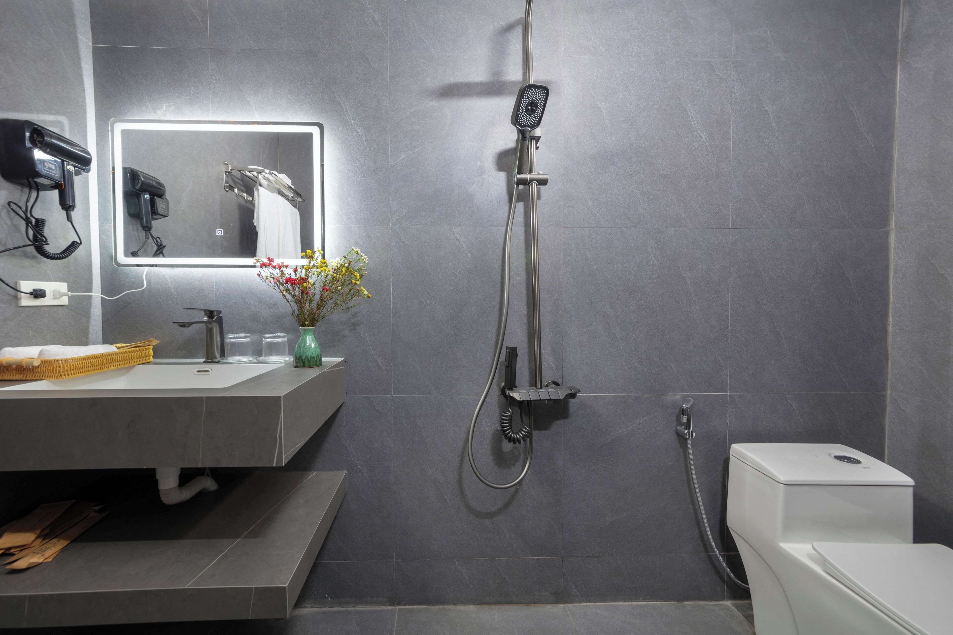 A modern bathroom with a grey tiled wall, a sink with a mirror, a handheld showerhead, and a toilet.
