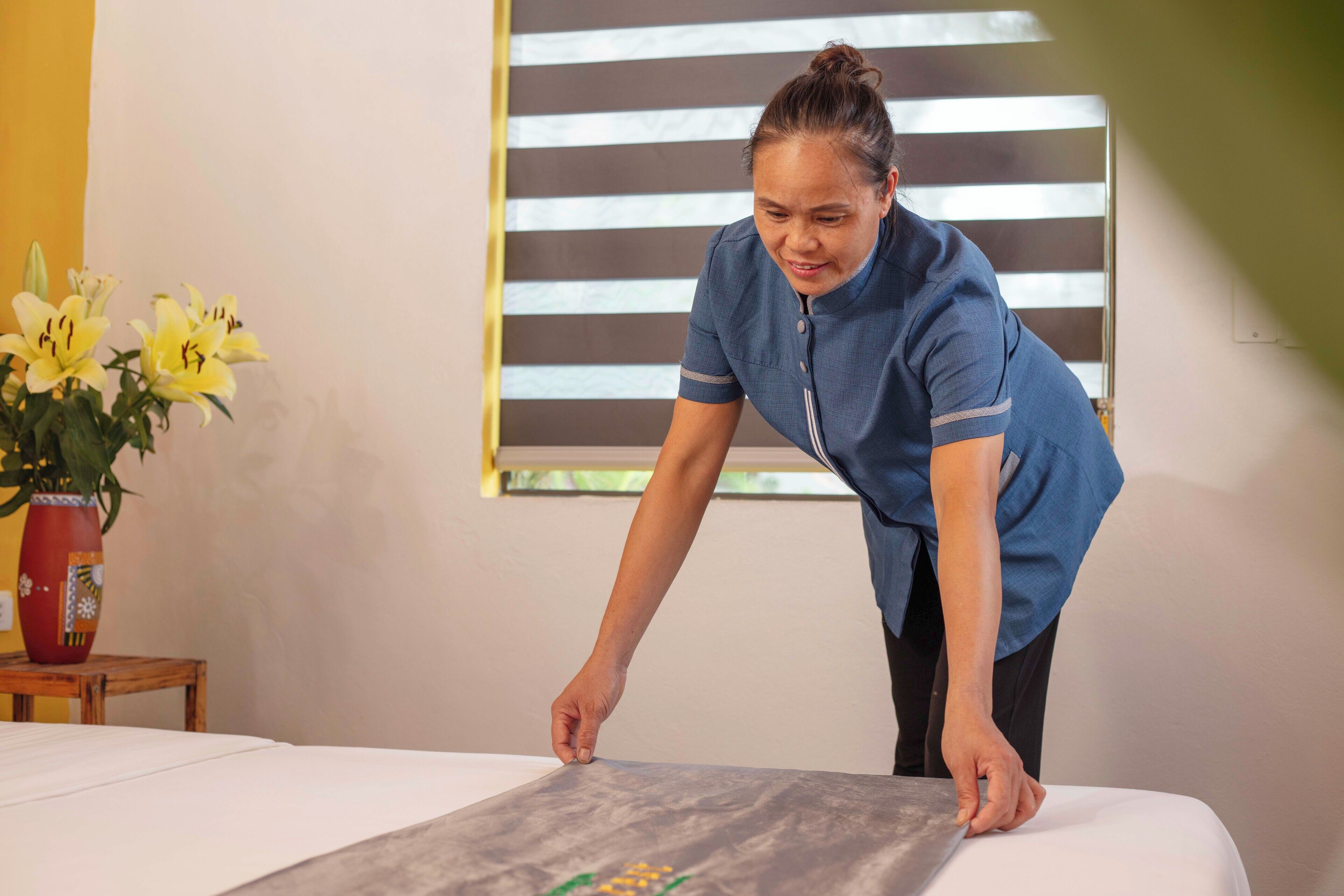 A person adjusting a bed in a room with a window, a vase with flowers, and a wall with a light switch.