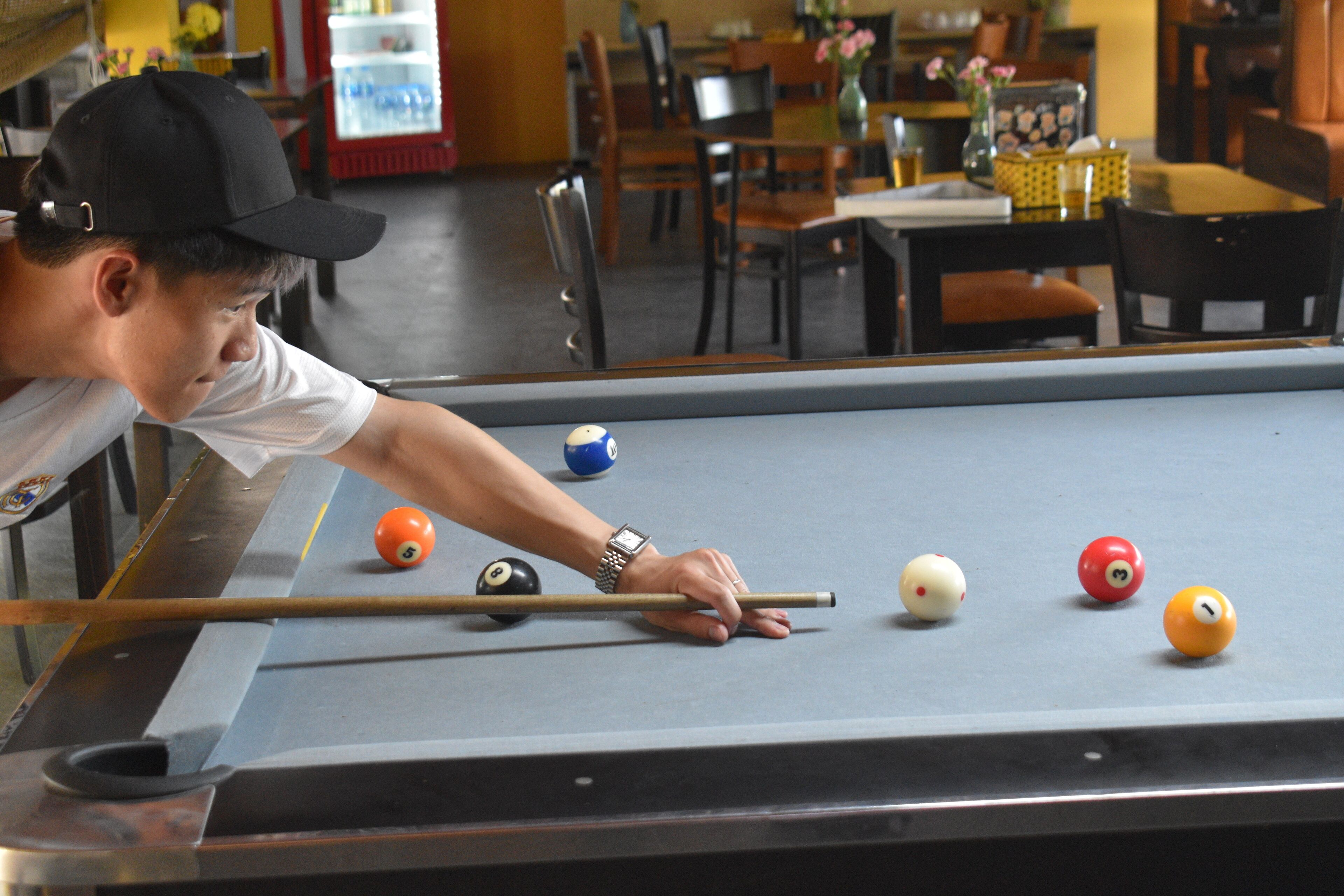 A pool table with a person playing, surrounded by chairs and tables in a room.