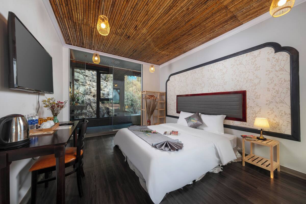 Habitación de hotel con una cama grande, un escritorio con hervidor, una silla, un televisor y una ventana con vistas a árboles.