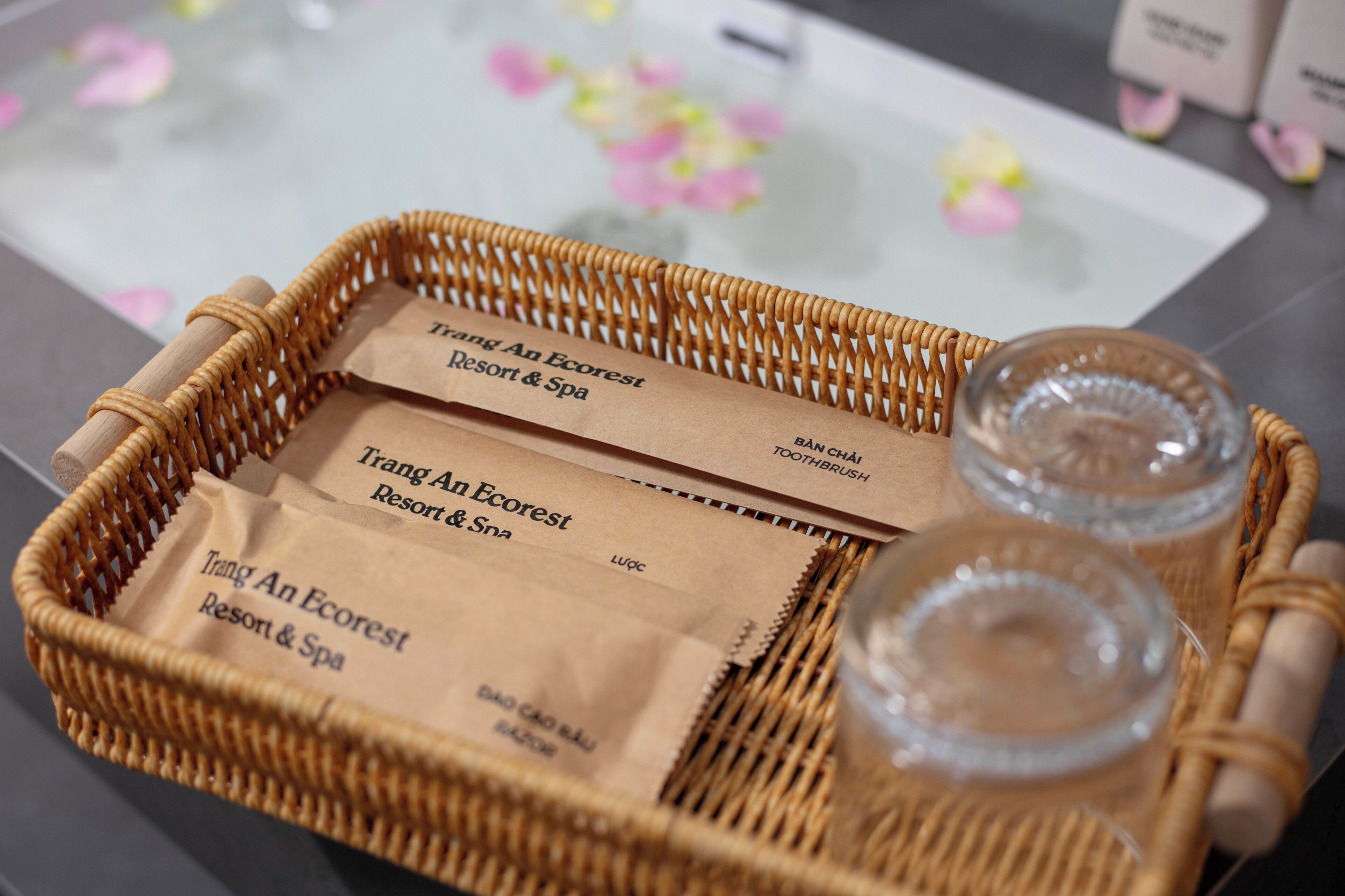 Uma bandeja de vime com dois potes de vidro e duas sacolas de papel pardo com a inscrição 'Trang An Ecorest Resort & Spa'.