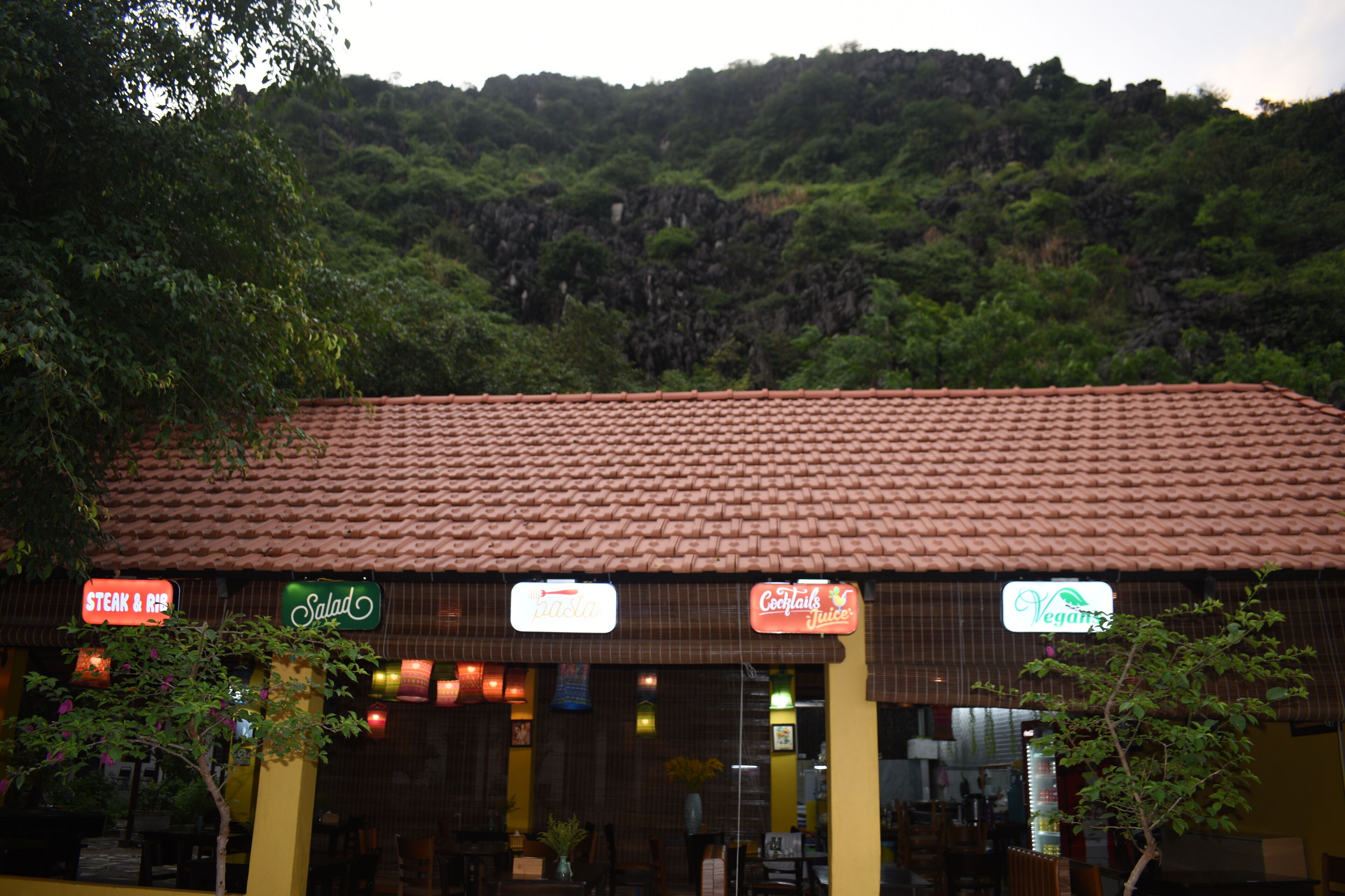 A restaurant with a tiled roof and signs for 'Steak & Rib', 'Salad', 'Pasta', 'Coconut Juice', and 'Vegan'.