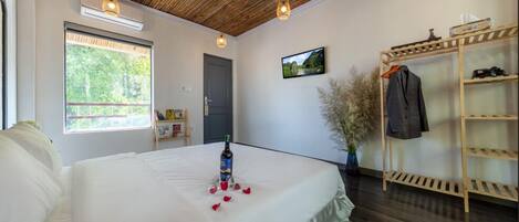 A bedroom with a large bed, a wooden ceiling, a wooden shelf with clothes, a TV, and a window with a view of greenery.