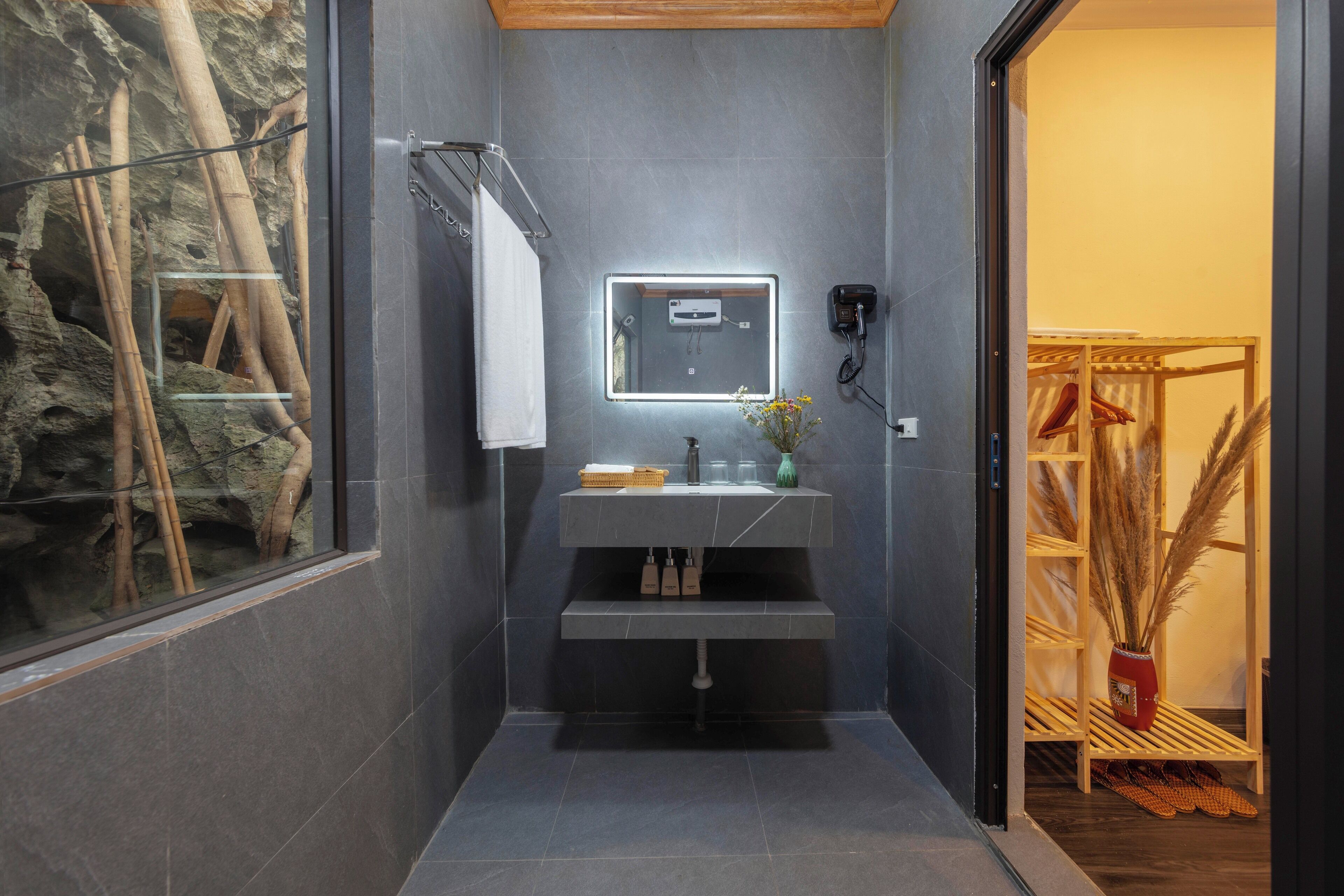 A modern bathroom with a dark tiled wall, a built-in sink, a mirror, and a window with a view of bamboo.