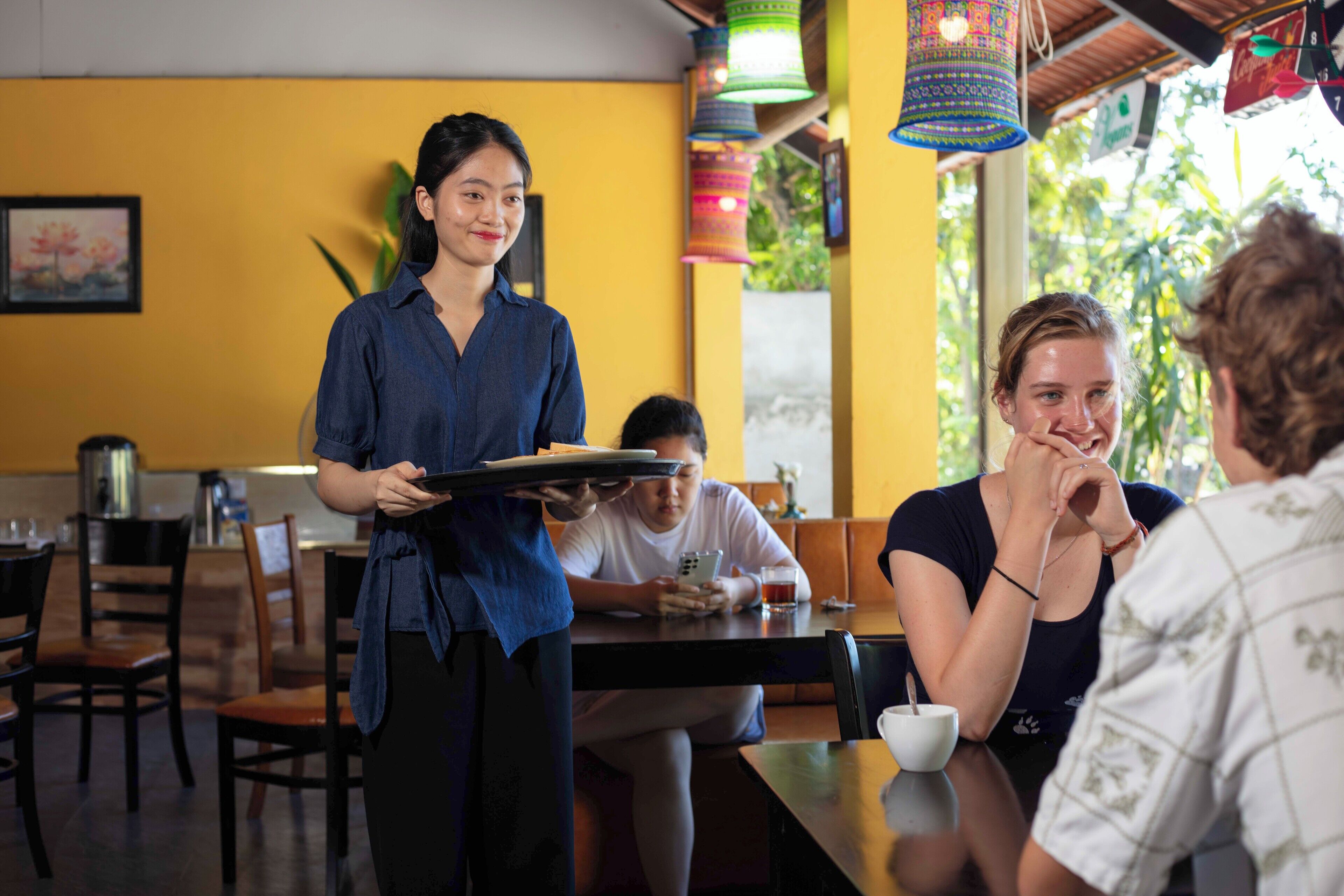 A restaurant with yellow walls, wooden tables, and colorful hanging lights.