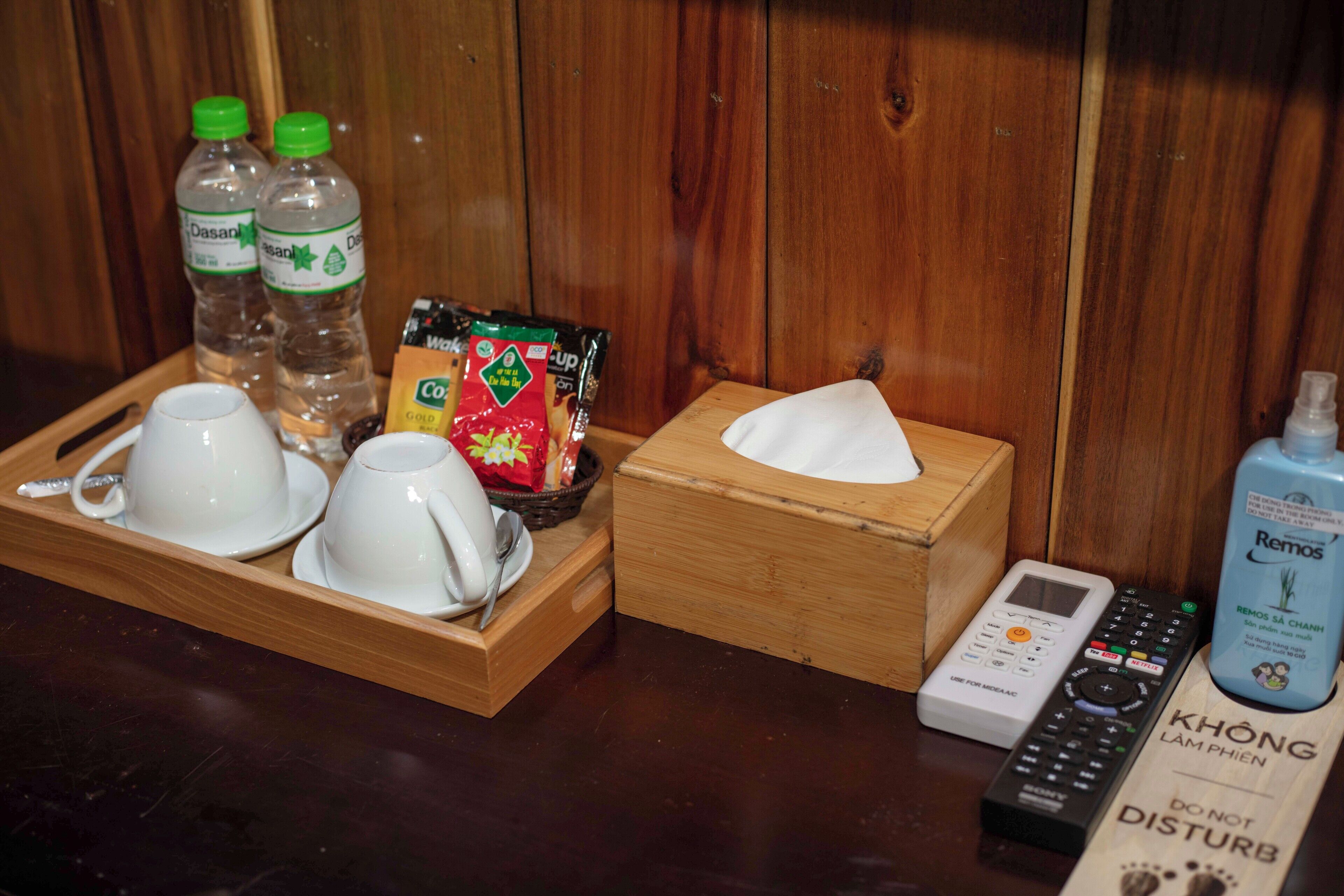 A wooden tray with white cups and saucers, Dasani water bottles, coffee packets, a remote control, and a tissue box.