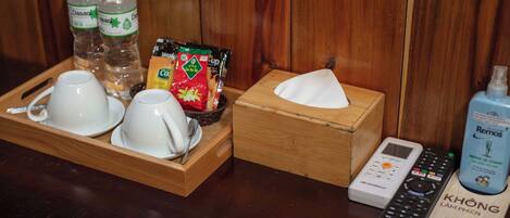 A wooden tray with white cups and saucers, Dasani water bottles, coffee packets, a remote control, and a tissue box.