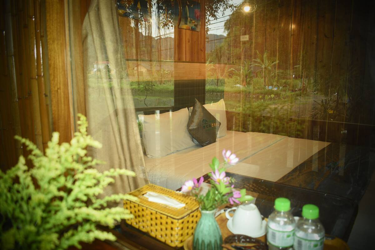 Una habitación con cama, ventana y una mesa con un florero y botellas.