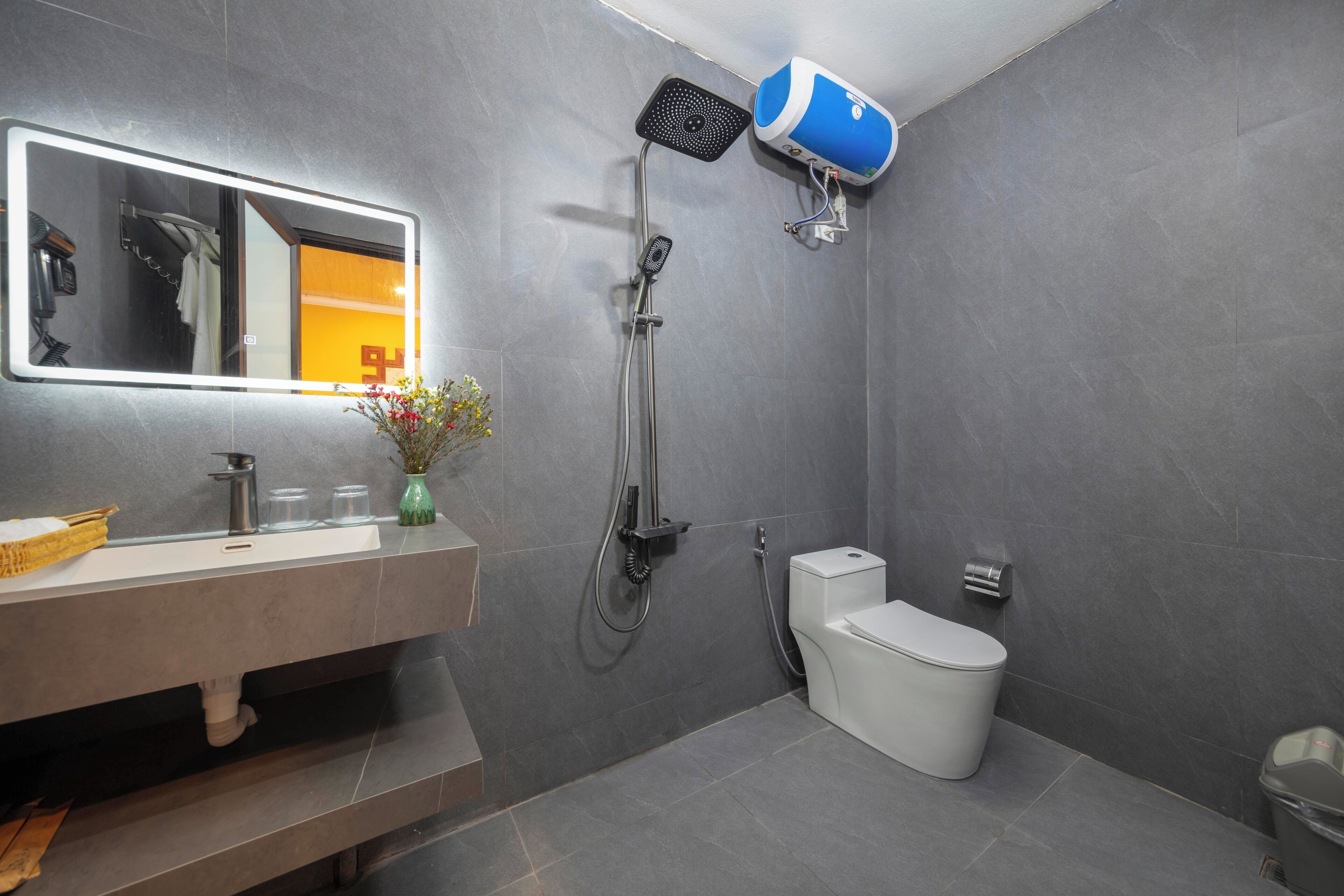 A modern bathroom with a large mirror, a showerhead, a toilet, and a sink with a vase of flowers.