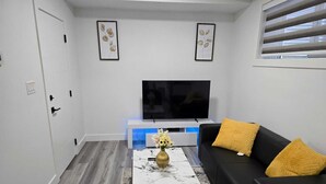 Un soggiorno moderno con una televisione a schermo piatto, un divano nero con cuscini gialli, un tavolino da caffè bianco con un vaso di fiori e un tappeto dal motivo marmorizzato.