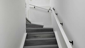 Una scala con gradini in legno e pareti bianche, che conduce a un pianerottolo con un quadro incorniciato.