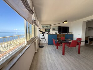 A modern apartment with a balcony overlooking a beach, a dining area with red chairs, and a kitchen with a TV. - Orizzonte Apartment in Follonica (Follonica)