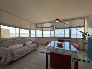 Een kamer met een glazen tafel, rode stoelen, een bank met bloemenprint en een telescoop op een statief.
