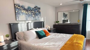 A modern hotel room with a large bed, a dark wooden nightstand, a bedside table with a lamp, a dresser, and a large mirror. - Your Perfect Home Away From Home · Owasso, OK (Owasso)