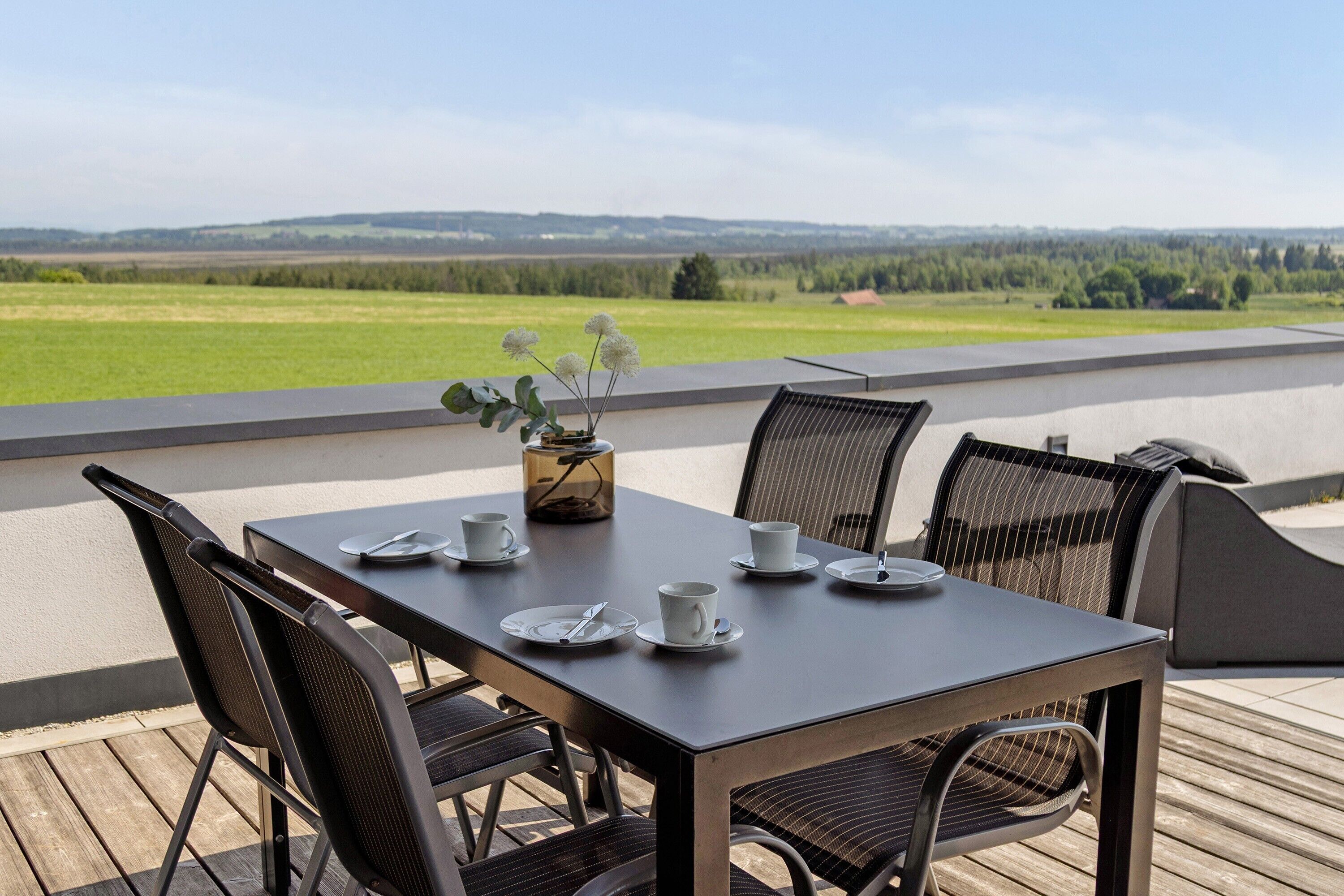 A table set outdoors with chairs, a vase with flowers, and a view of a green landscape.