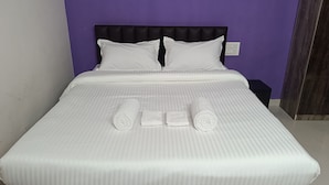 A neatly made bed with white linens and two pillows, a rolled towel, and two folded towels on a bed in a room with purple walls. - Kokan Inn (Dapoli)