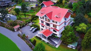 Un edificio de varios pisos con techo rojo, rodeado de abundante vegetación y un pequeño estacionamiento.