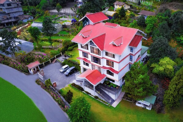 Residential building with a red roof, surrounded by lush greenery and a well-maintained garden, offering multiple balconies and ample parking space.