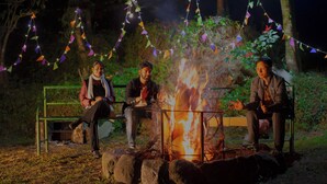 Tres personas sentadas alrededor de una fogata con luces de colores al fondo.