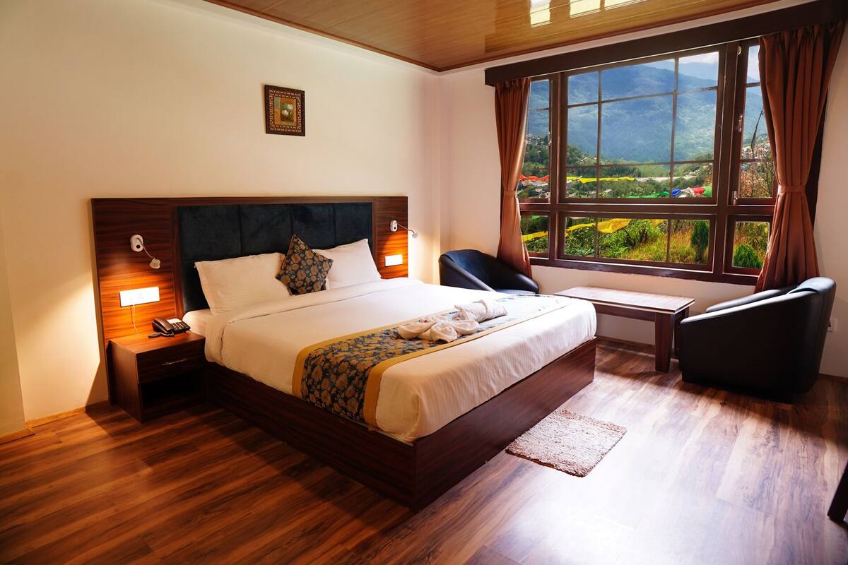 Habitación de hotel con una cama grande, mesitas de noche, un escritorio y vistas a un paisaje montañoso a través de un ventanal amplio.