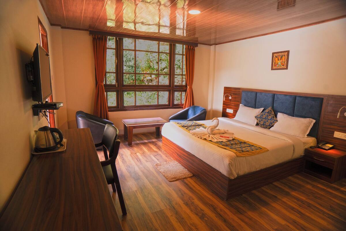 Habitación de hotel con suelo de madera, cama con cabecera azul, escritorio con silla, ventana con cortinas y televisor montado en la pared.