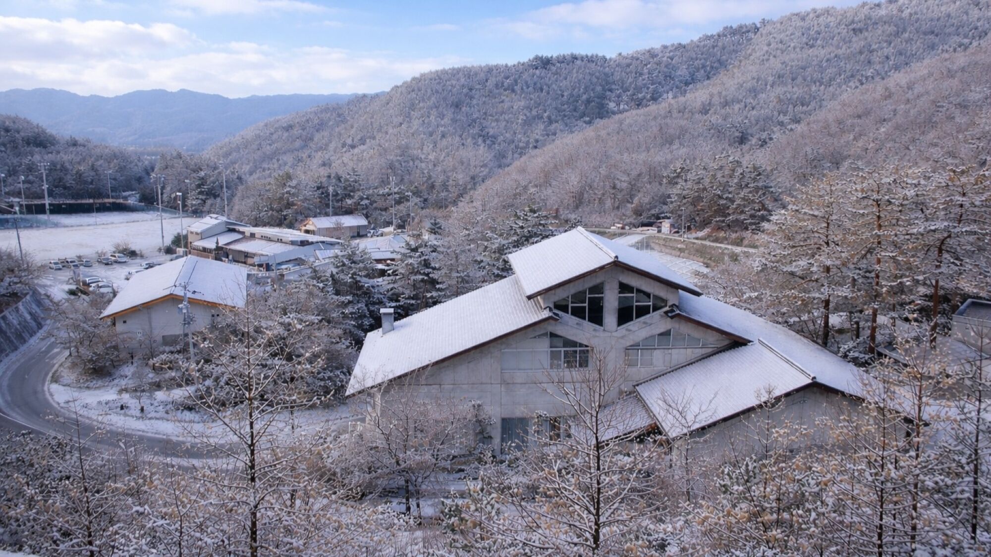 A building with a sloped roof and large windows, surrounded by snow-covered trees and a mountainous backdrop.