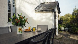 Ein Esstisch ist gedeckt mit Frühstücksgebäck, ein Blumenstrauß steht darauf und im Hintergrund ist ein weißes Gebäude mit geneigtem Dach zu sehen.