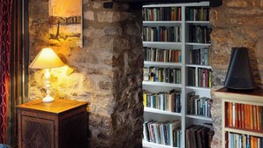Ein gemütliches Zimmer mit einer Steinswand, einem Bücherregal voller Bücher, einer Lampe auf einem Holznachtisch und einem Gemälde an der Wand.