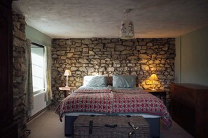 A bedroom with a stone wall, a bed with patterned bedding, two bedside lamps, and a wooden chest of drawers. - Large Country House | Games Room & Log Burner (Haverfordwest)