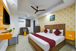 A hotel room with a large bed, a ceiling fan, a TV mounted on the wall, a small fridge, and a bedside table with a plant. - The Kelvish Plaza (New Delhi)