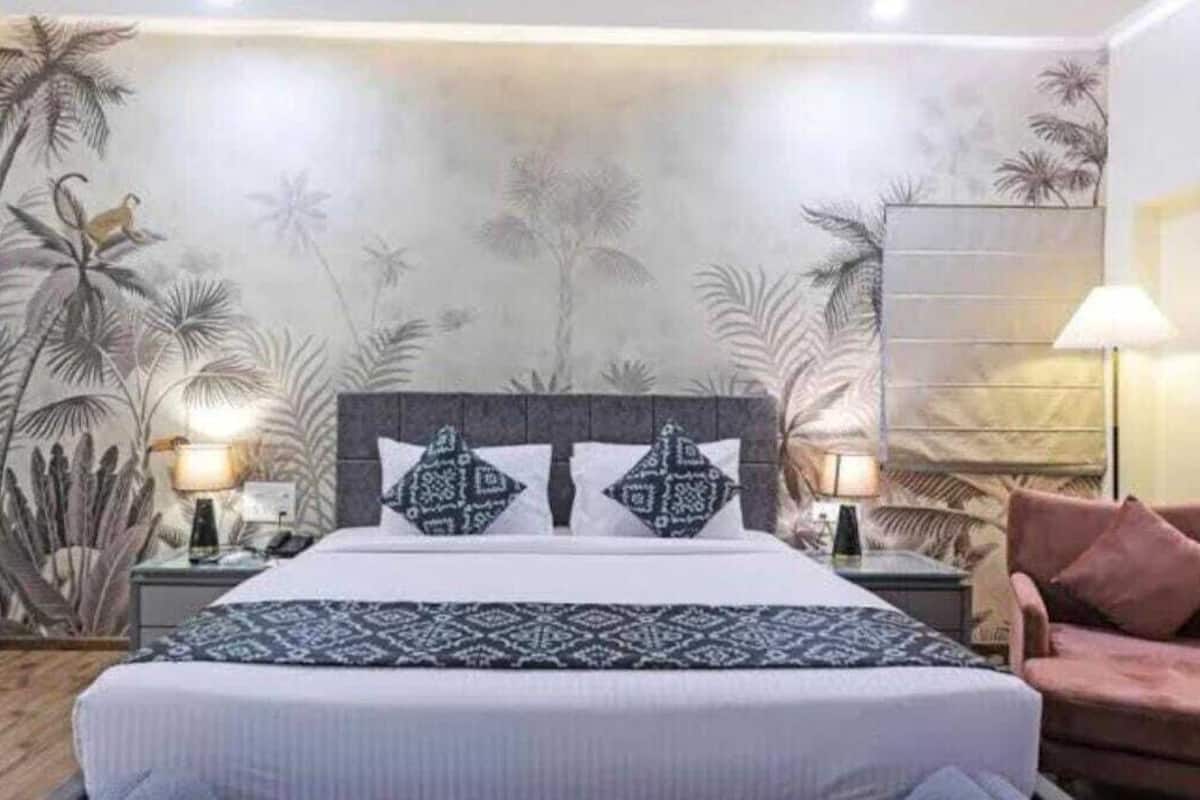 Un dormitori amb un llit, dues taules de nit, una làmpada, una cadira i una paret amb un mural tropical.