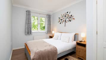 A bedroom with a bed, two bedside tables, a window with curtains, a radiator, and a wall-mounted decorative piece.