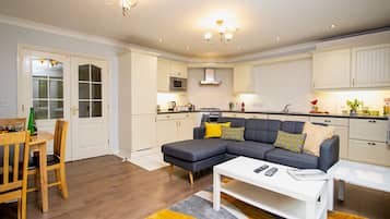A modern living room with a sectional sofa, a dining area with a wooden table and chairs, and a kitchen with white cabinets and a microwave.