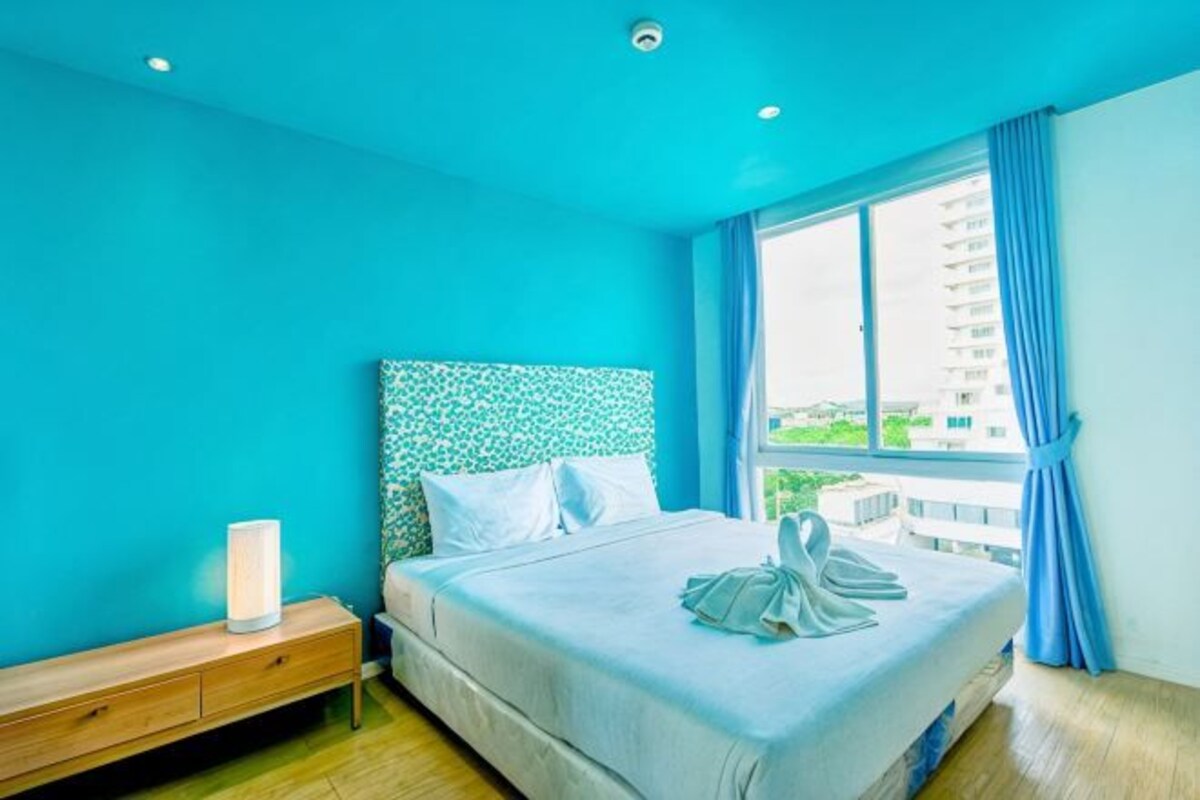 A hotel room with a large bed, a nightstand, a window with a view of buildings, and a blue wall.