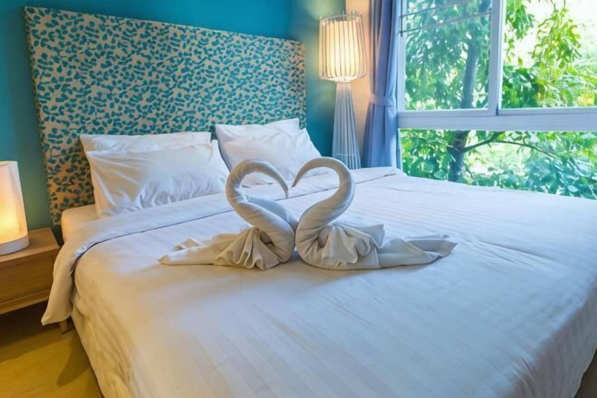 Een bed met een handdoekopstelling in de vorm van een hart, een gestreept hoofdeinde en een raam met uitzicht op bomen.