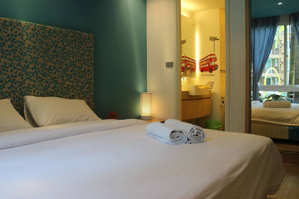 Een hotelkamer met een bed, nachtkastje, lamp, spiegel en uitzicht op de straat door een raam.