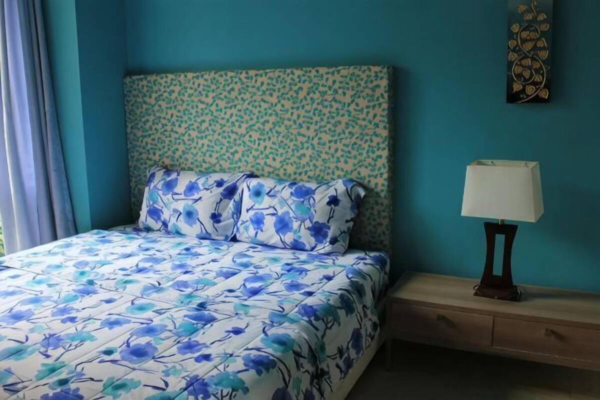 Een bed met bloemenbeddengoed, een nachtkastje met een lamp, en een kunstwerk aan de muur.