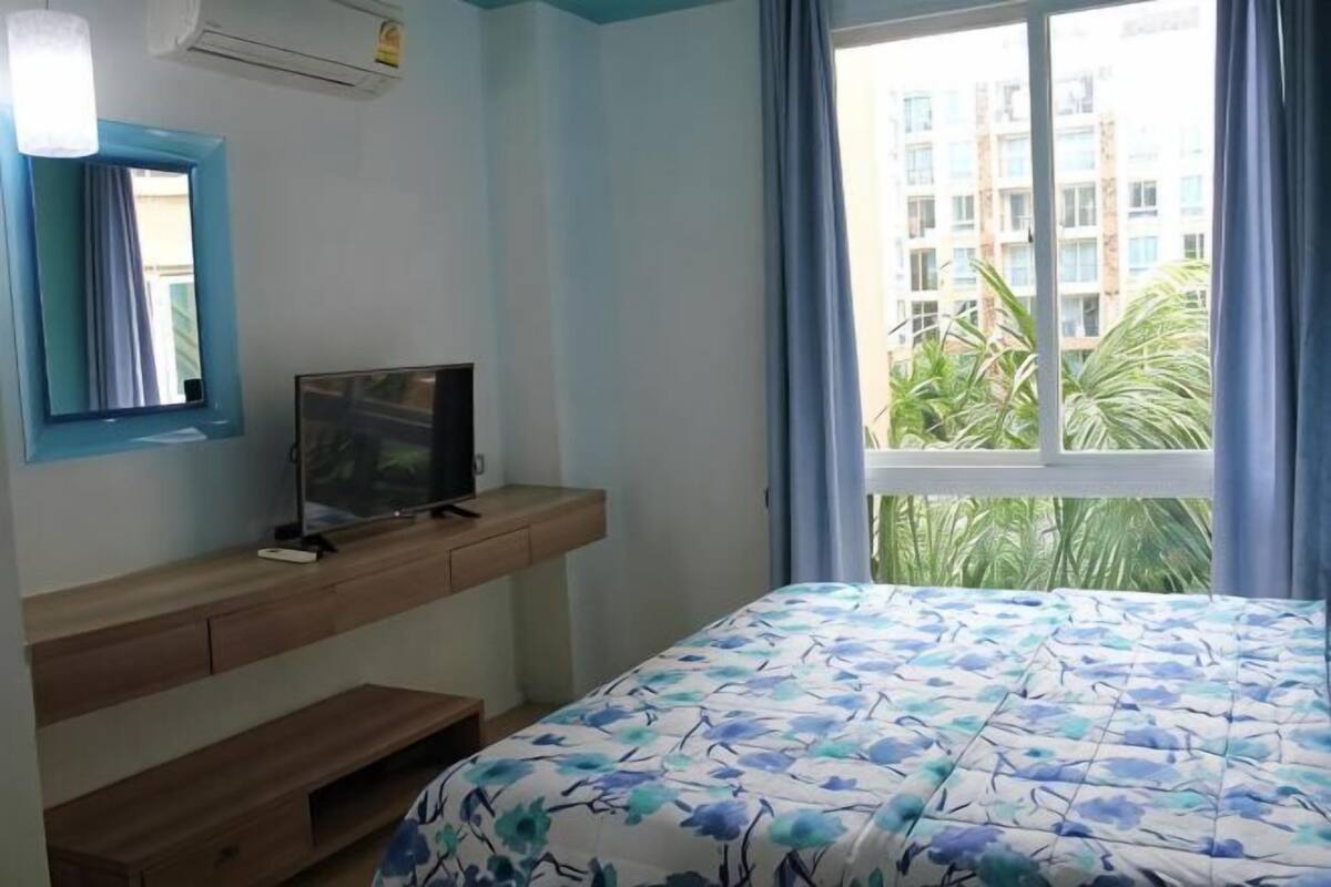 Een slaapkamer met een bed, een televisie op een houten standaard, een raam met gordijnen en uitzicht op gebouwen en planten.