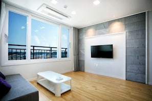 A modern living room with large windows, a flat-screen TV, a white sofa, and a wooden floor.